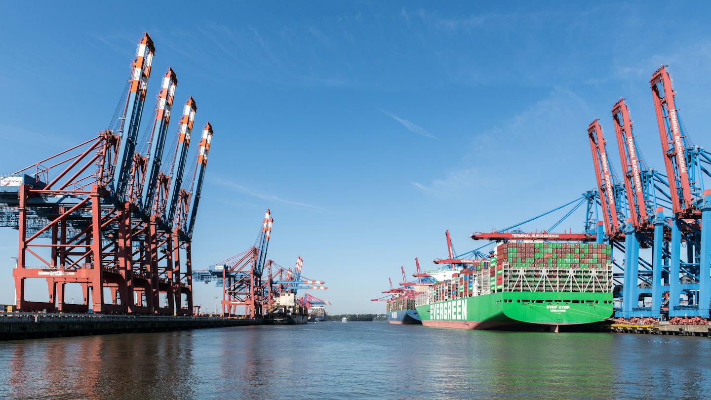 A large green container ship is docked at a modern port with cranes.